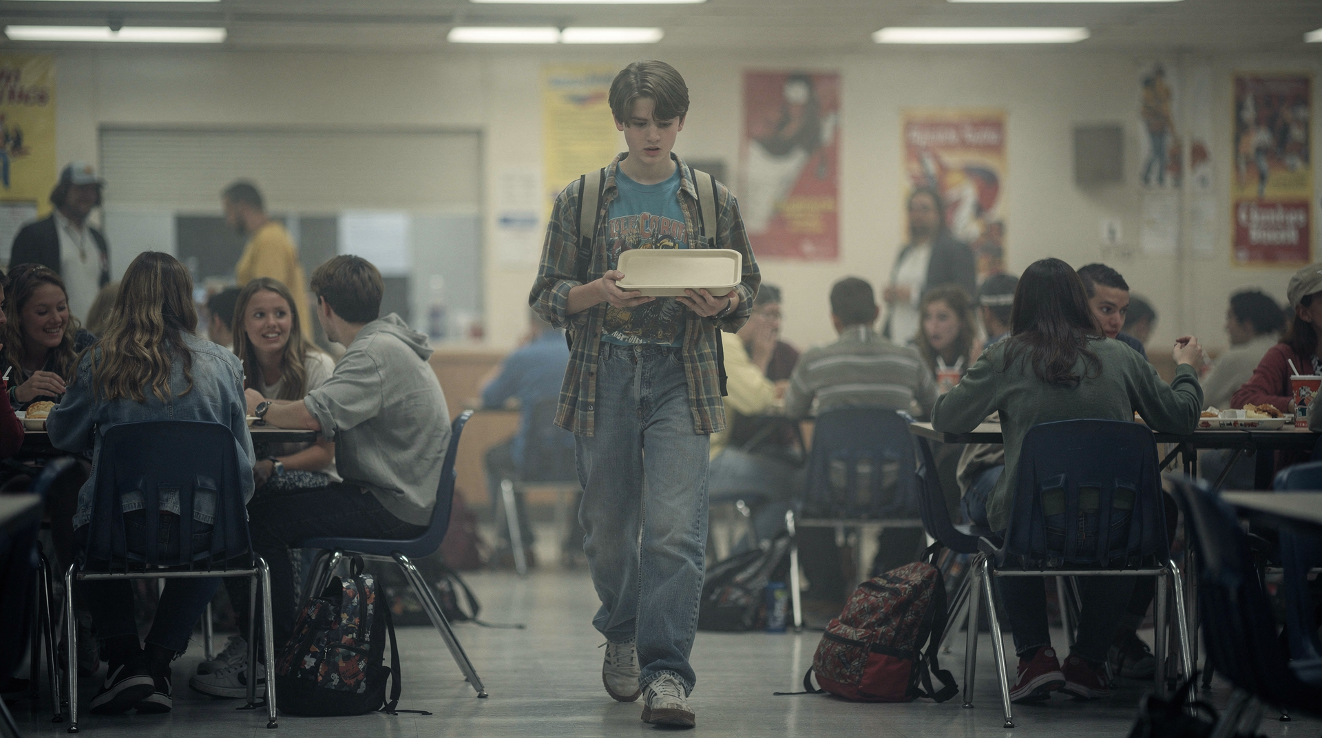 A teenage boy walking alone through a high school cafeteria with a lunch tray, eyes down, surrounded by full tables